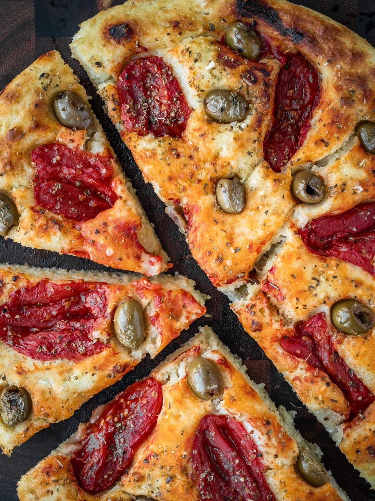 Focaccia Barese slices garnished with tomatoes, green olives and oregano on a wooden board.