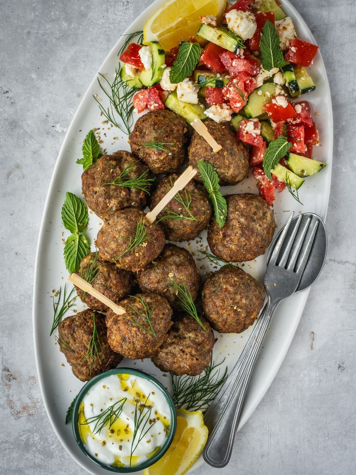 Greek Meatballs on a platter garnished with fresh mint and dill with a side of Greek salad and yogurt dip.
