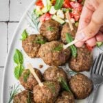 A hand picking up a Greek Meatball from a platter garnished with fresh mint and dill, with a side of Greek salad and yogurt dip.