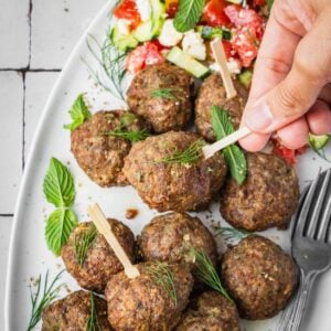 A hand picking up a Greek Meatball from a platter garnished with fresh mint and dill, with a side of Greek salad and yogurt dip.