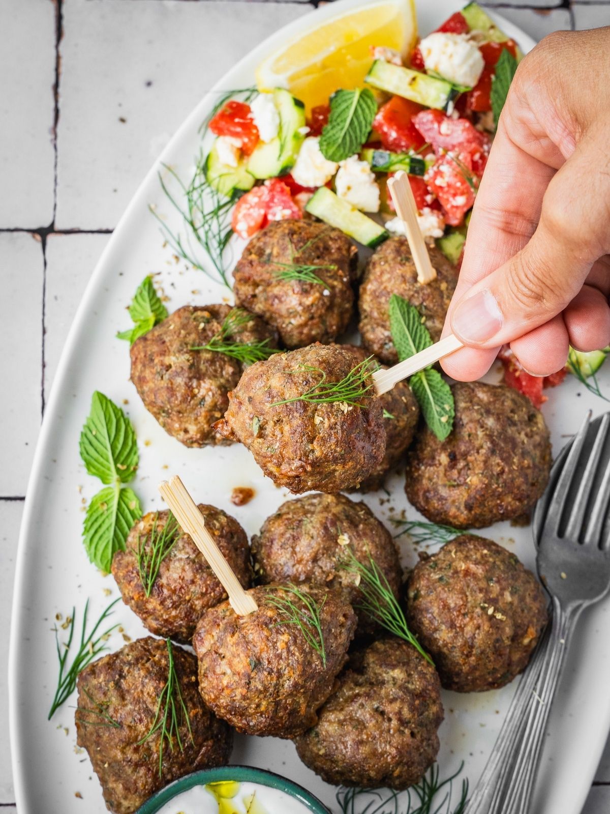 A hand picking up a Greek Meatball from a platter garnished with fresh mint and dill, with a side of Greek salad and yogurt dip.