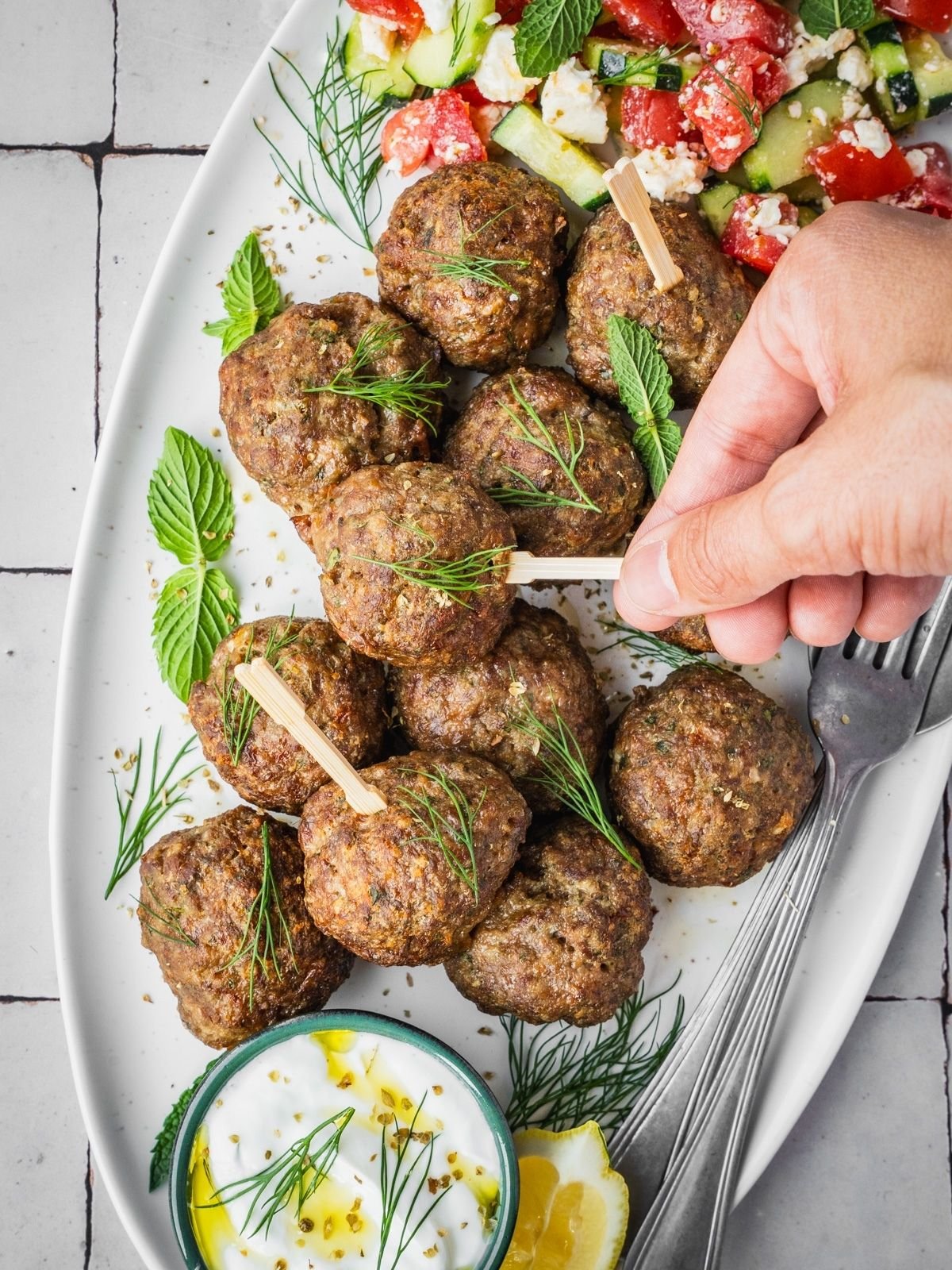 A hand picking up a Greek Meatball from a platter garnished with fresh mint and dill, with a side of Greek salad and yogurt dip.