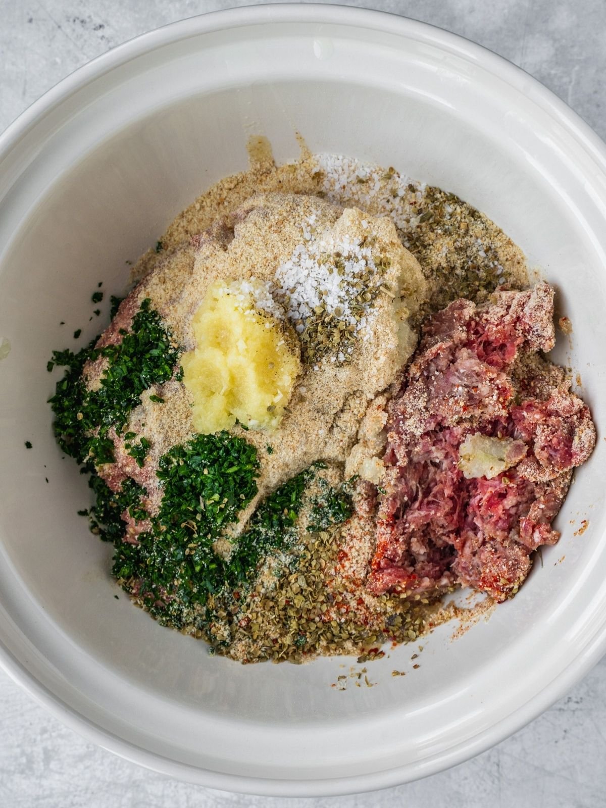 A large mixing bowl with ground beef, egg, breadcrumbs, herbs, spices, onion and garlic.