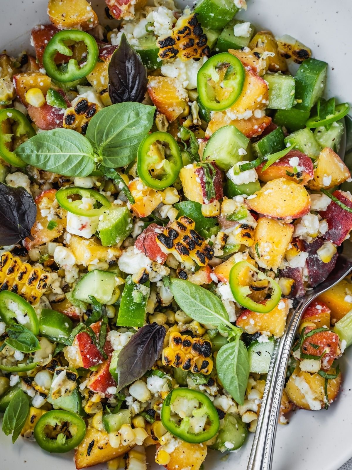 Grilled corn and peach summer salad with chopped cucumber and crumbled feta in a bowl garnished with jalapeno pepper rings and basil leaves.