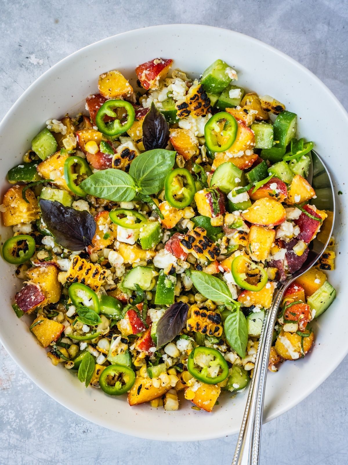 Grilled corn and peach summer salad with chopped cucumber and crumbled feta in a bowl garnished with jalapeno pepper rings and basil leaves.