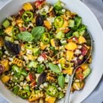 Grilled corn and peach summer salad with chopped cucumber and crumbled feta in a bowl garnished with jalapeno pepper rings and basil leaves.