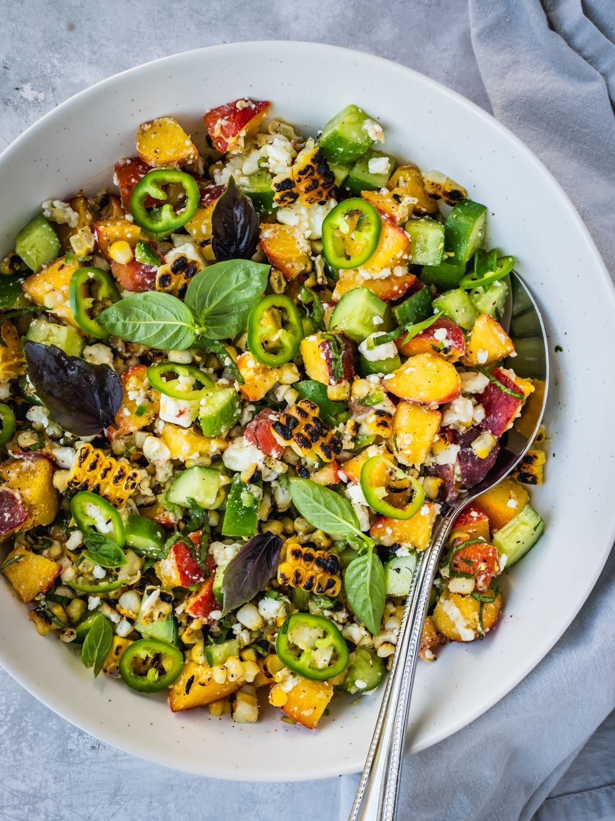 Grilled corn and peach summer salad with chopped cucumber and crumbled feta in a bowl garnished with jalapeno pepper rings and basil leaves.