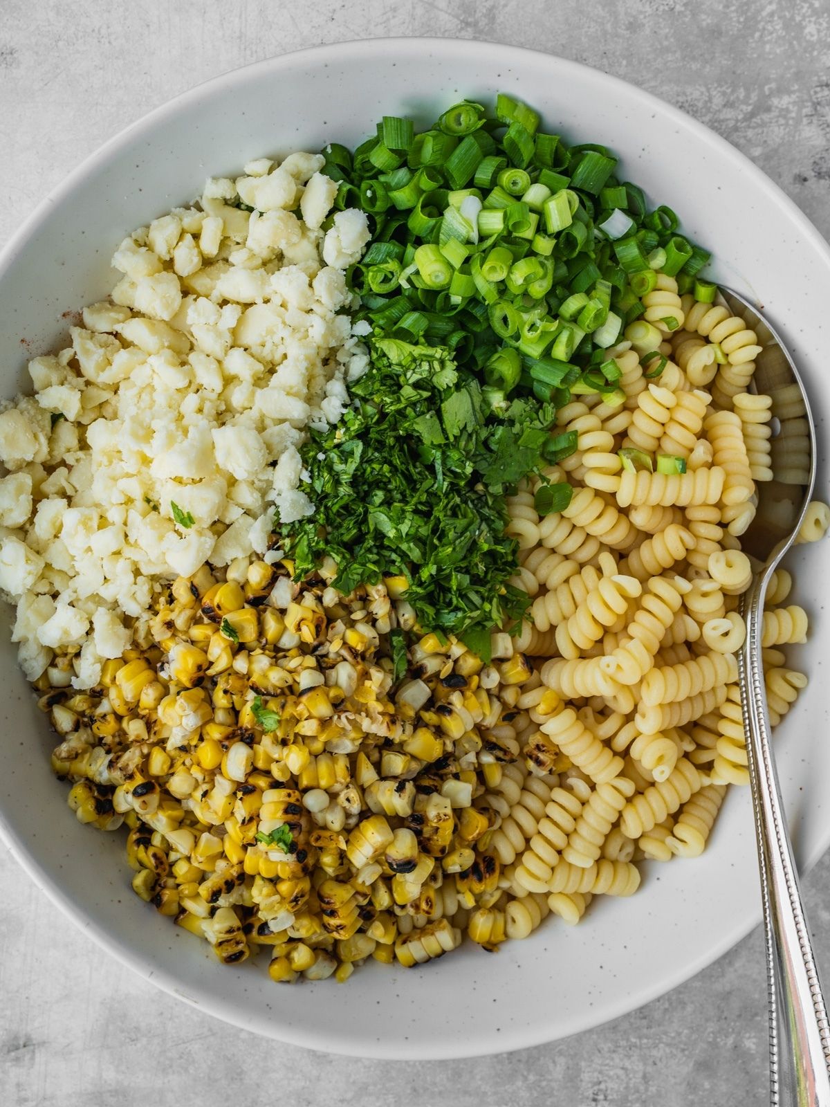 Cooked pasta, corn kernels, chopped green onion, chopped cilantro and crumbled cotija cheese in the serving bowl.