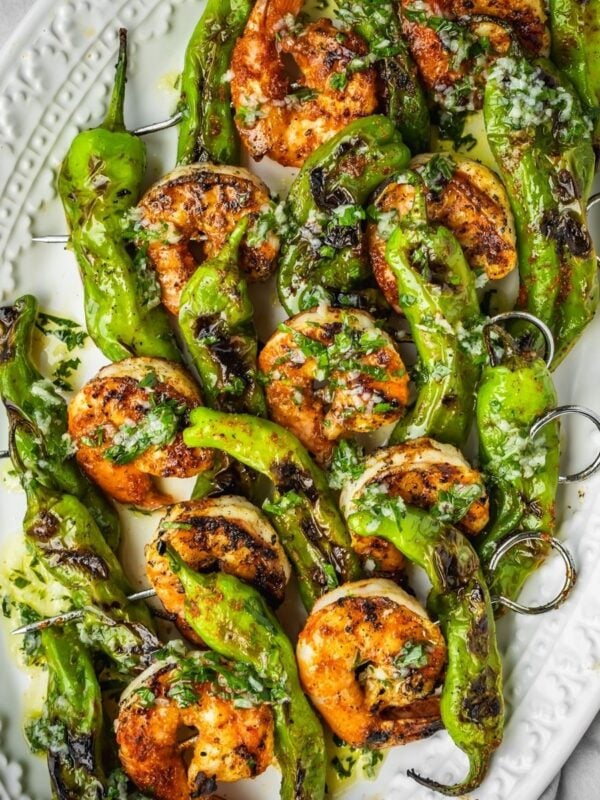 Grilled shrimp and shishito pepper skewers on a platter garnished with garlic and herb oil.