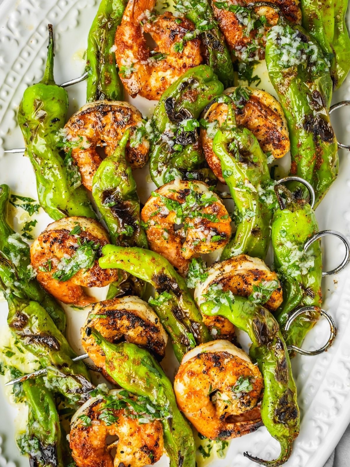 Grilled shrimp and shishito pepper skewers on a platter garnished with garlic and herb oil.