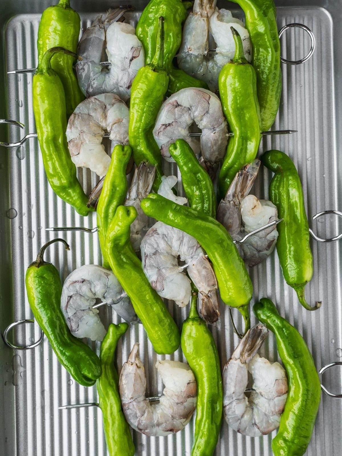 Raw prawns and shishito peppers on metal skewers.