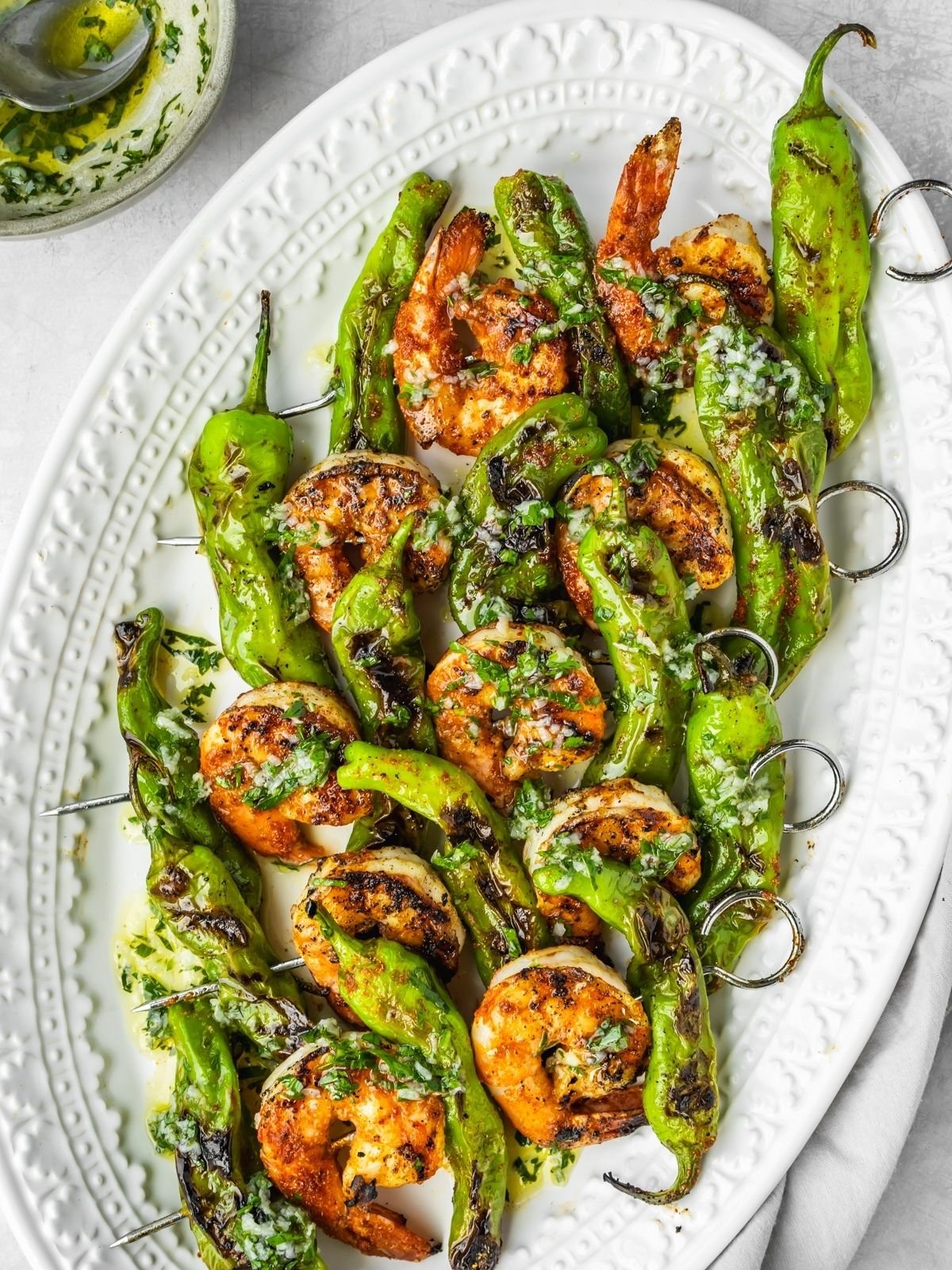 Grilled prawns and shishito pepper skewers on a platter garnished with garlic and herb oil.