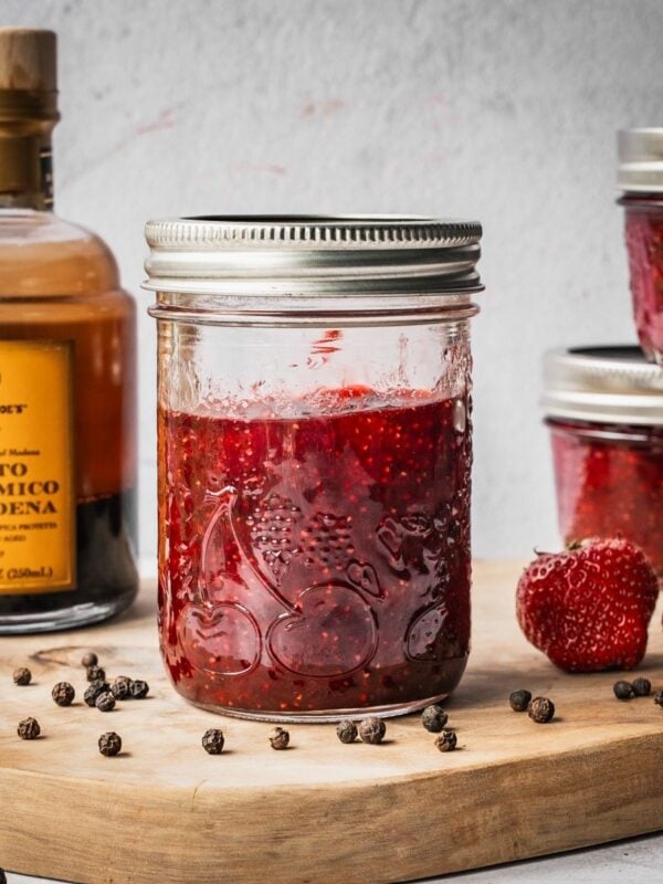Strawberry Balsamic Black Pepper Jam in mason jars on a wooden board with fresh strawberries, black peppercorns and a bottle of balsamic vinegar on the side.