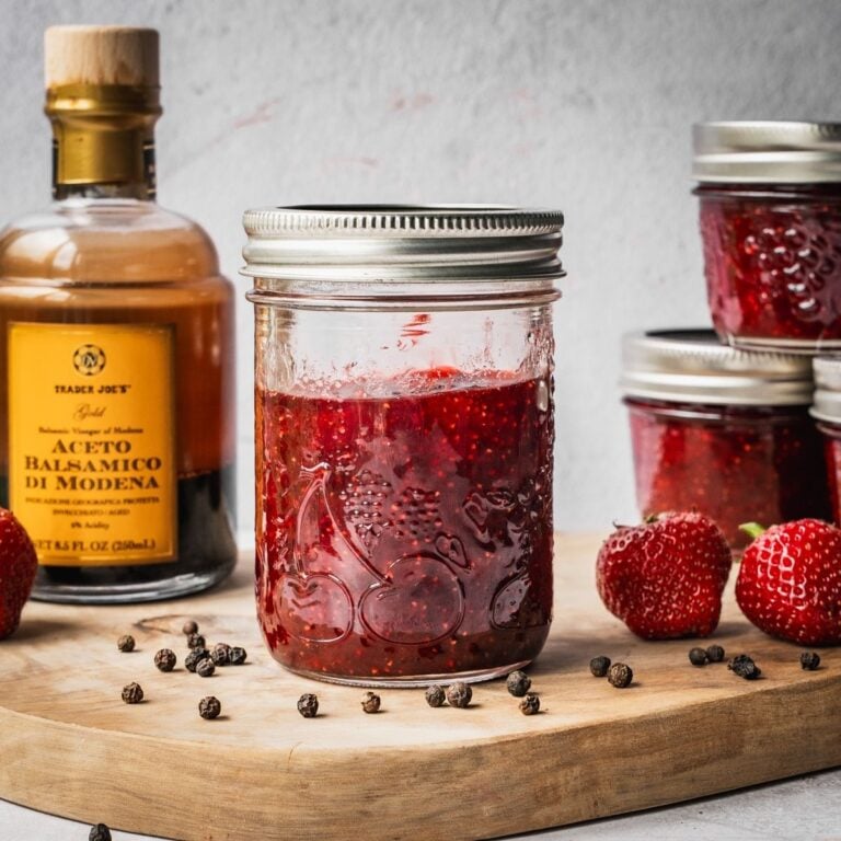Strawberry Balsamic Black Pepper Jam in mason jars on a wooden board with fresh strawberries, black peppercorns and a bottle of balsamic vinegar on the side.