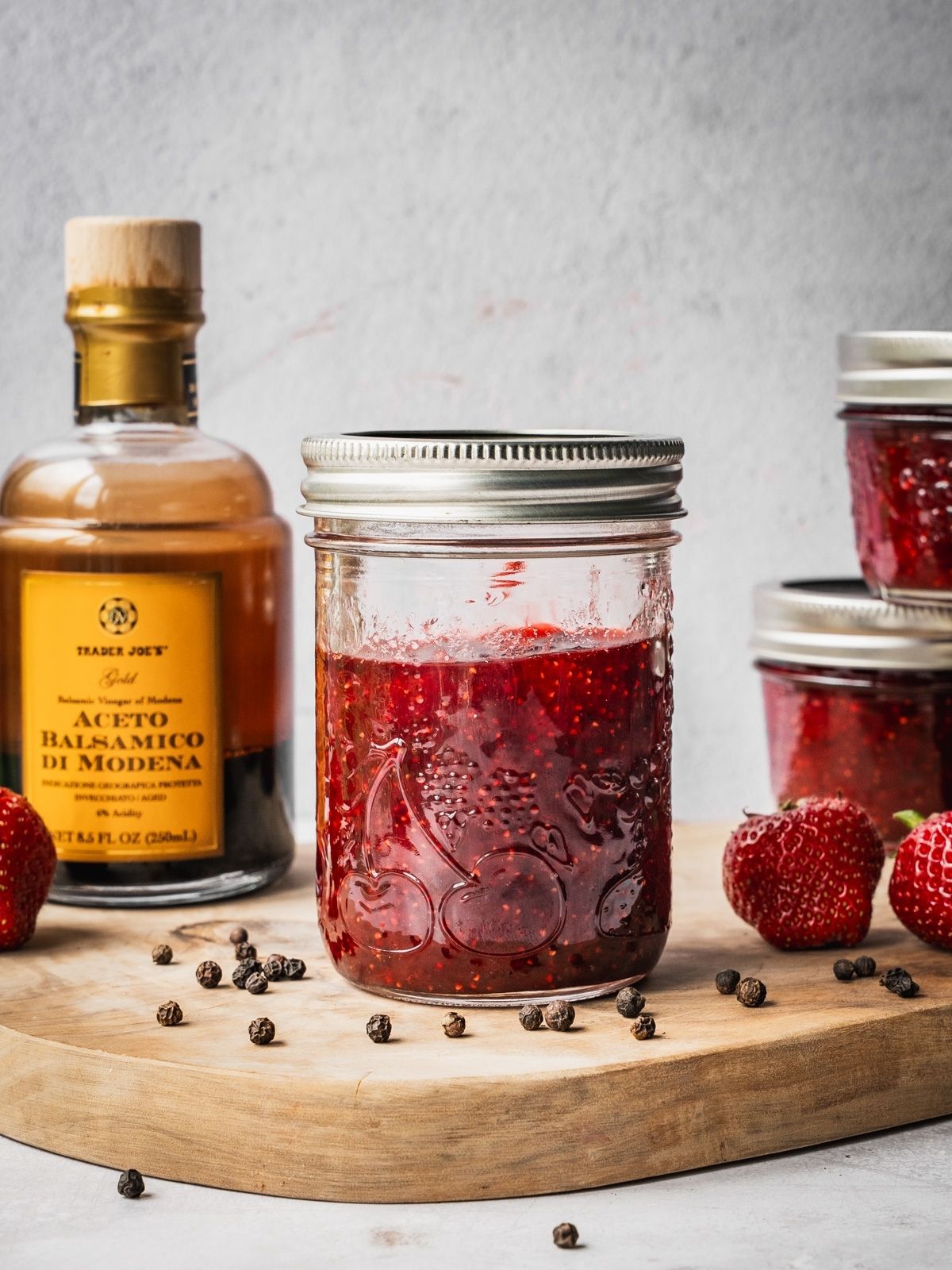 Strawberry Balsamic Black Pepper Jam in mason jars on a wooden board with fresh strawberries, black peppercorns and a bottle of balsamic vinegar on the side.
