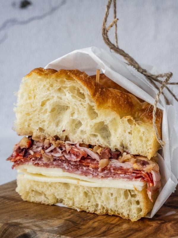 Muffuletta sandwich made with fresh homemade focaccia stuffed with cheese and deli meat, wrapped in paper and sitting on a wooden tray.