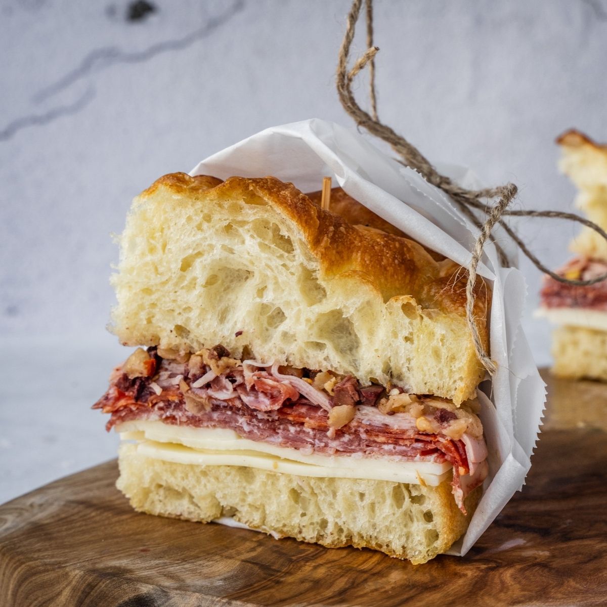 Muffuletta sandwich made with fresh homemade focaccia stuffed with cheese and deli meat, wrapped in paper and sitting on a wooden tray.