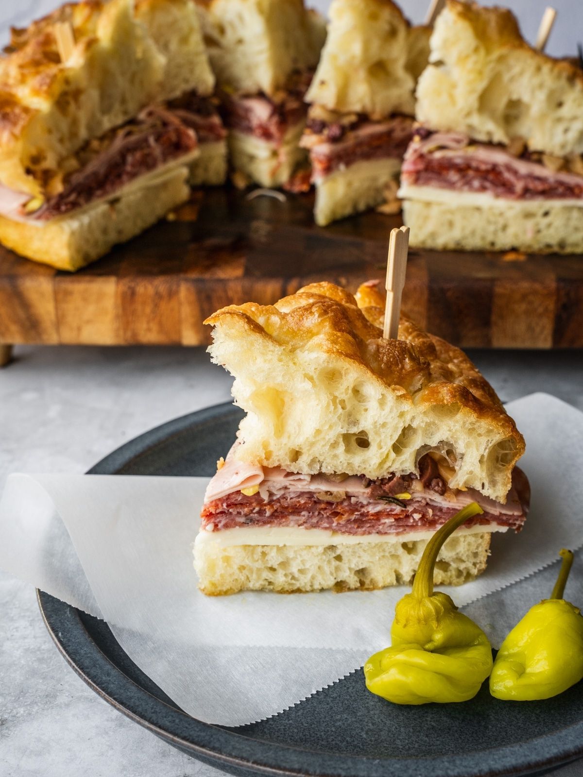 Muffuletta sandwich made with fresh homemade focaccia stuffed with cheese and deli meat on a plate with a side of hot peppers.