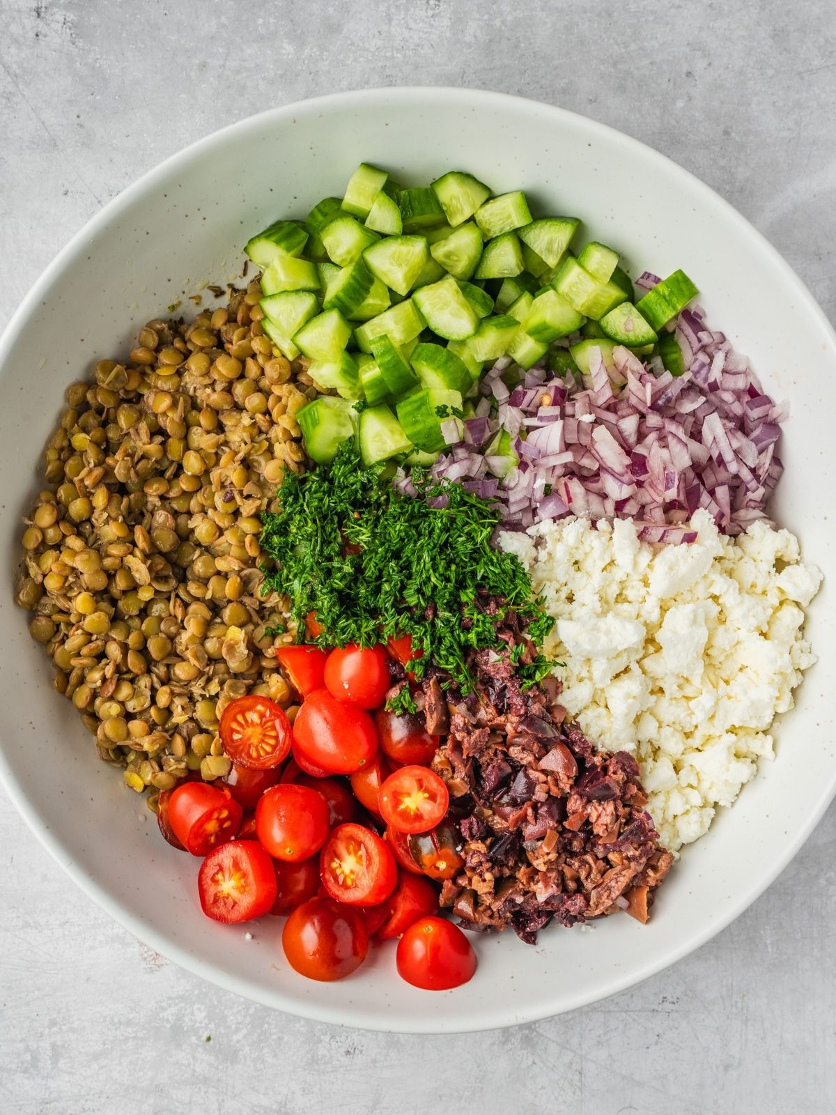 Large bowl with cooked lentils, chopped cucumbers, sliced cherry tomatoes, crumbled feta, chopped olives and herbs.