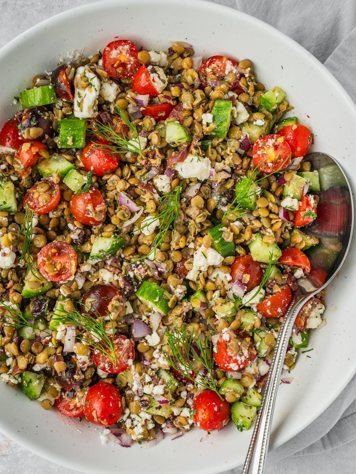 Greek lentil salad in a large bowl with sliced cherry tomatoes, cucumbers, feta, olives and herbs.