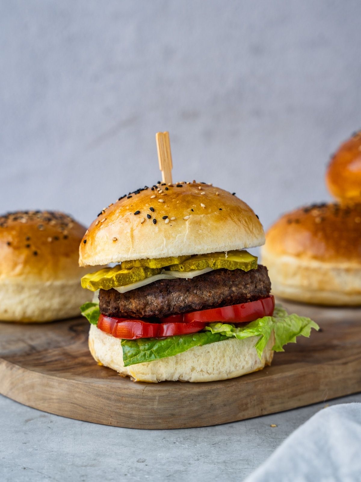Hamburger bun stuffed with a burger patty and vegetables on a wooden tray with more buns in the background.