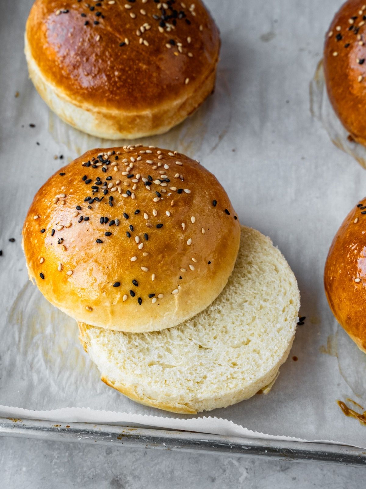Hamburger buns on a parchment paper-lined baking sheet, with one cut exposing the tight crumb.