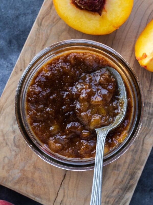 Maple peach chutney in a mason jar on a wooden board with fresh peaches on the side.