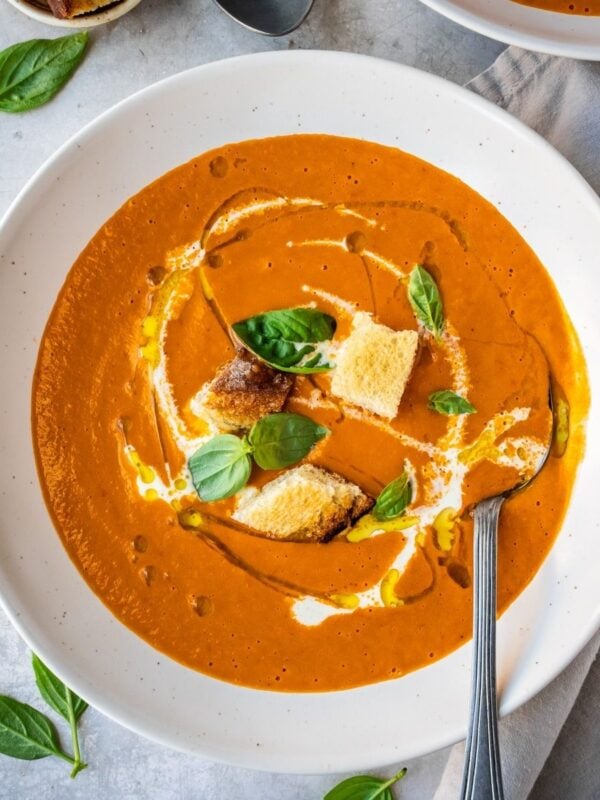 Roasted red pepper and tomato soup in a bowl, garnished with croutons, fresh basil, olive oil and cream.