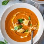 Roasted red pepper and tomato soup in a bowl, garnished with croutons, fresh basil, olive oil and cream.