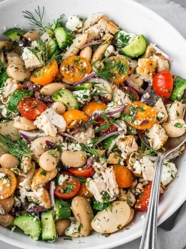 Bowl of bean salad with chicken pieces, cherry tomatoes, cucumbers and herbs.