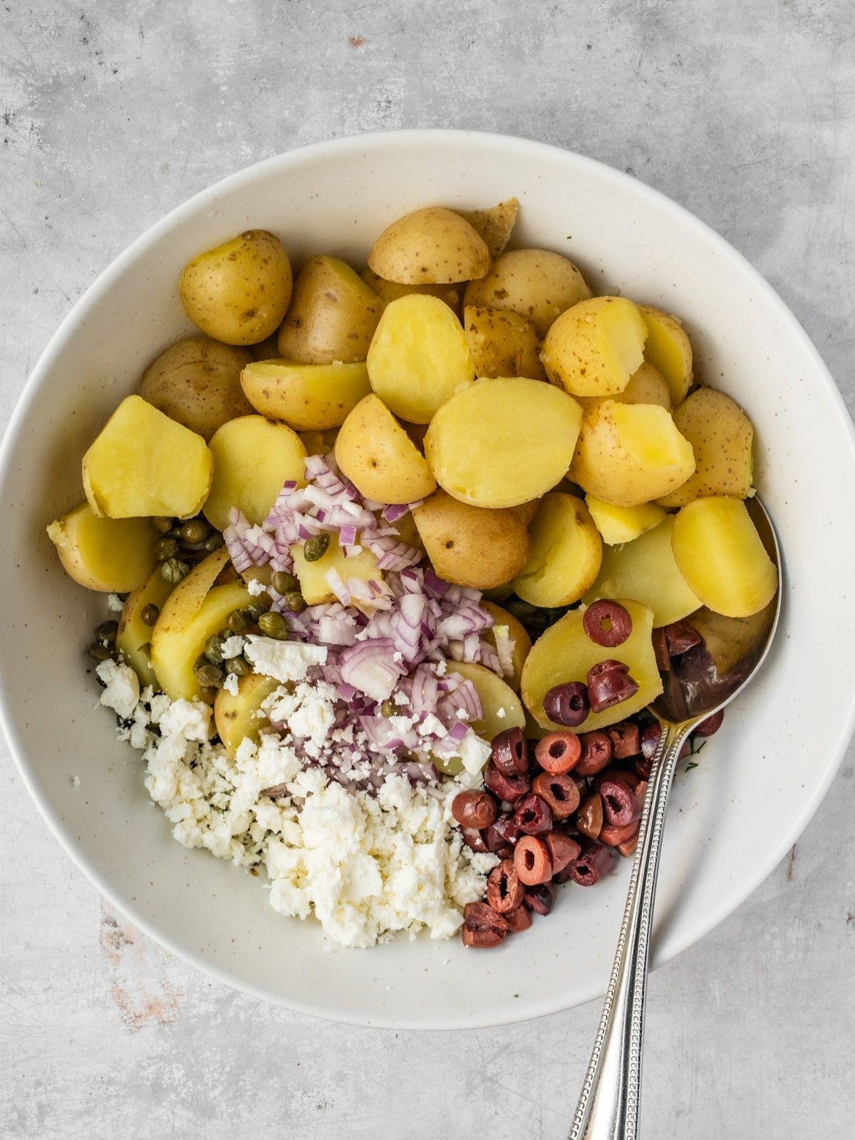 Large serving bowl with cooked and cooled potato chunks, chopped olives, capers, and feta.