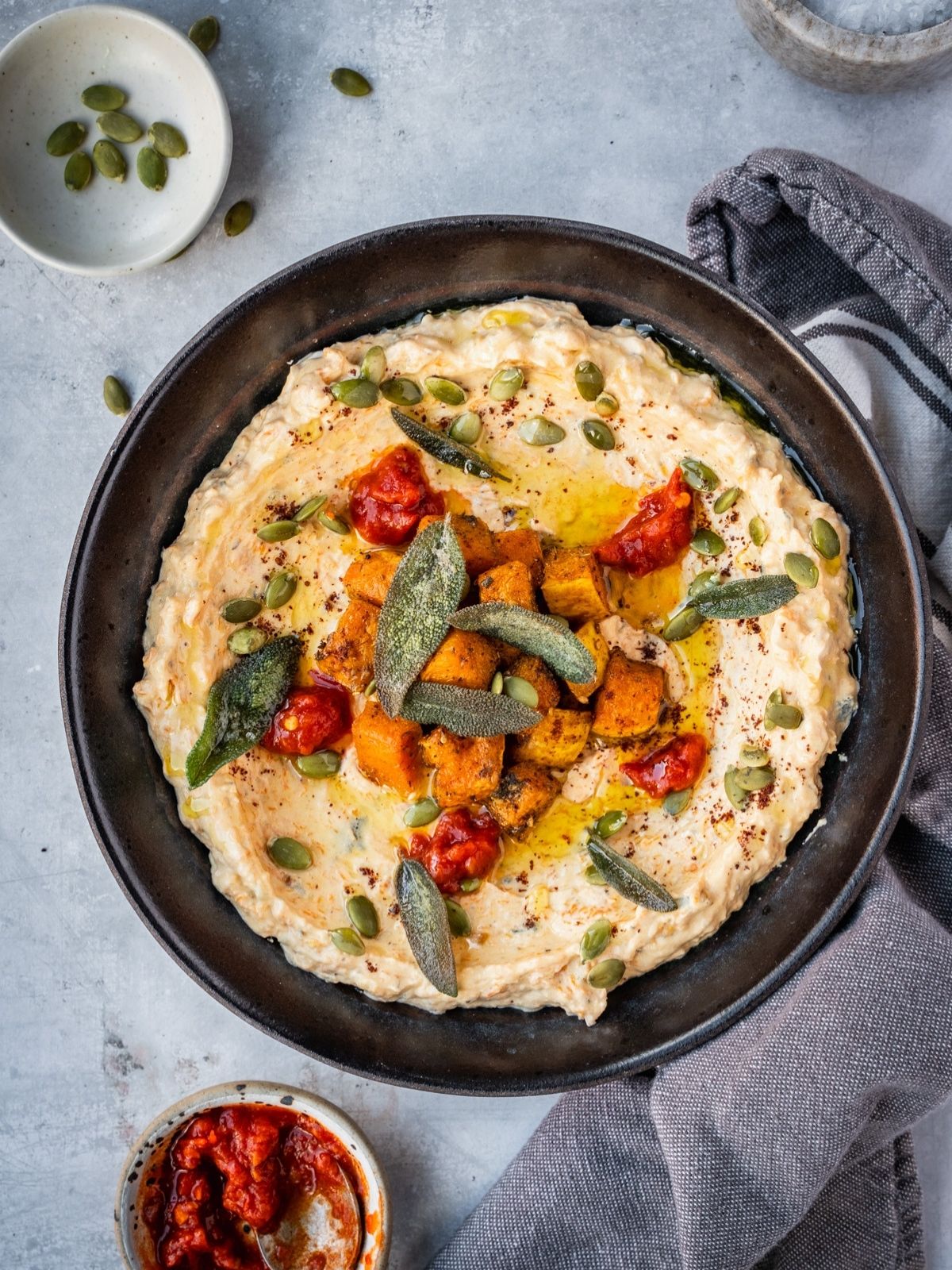A bowl with mafghoussa sip garnished with roasted squash, pumpkin seeds, fried sage, sumac and chili paste.