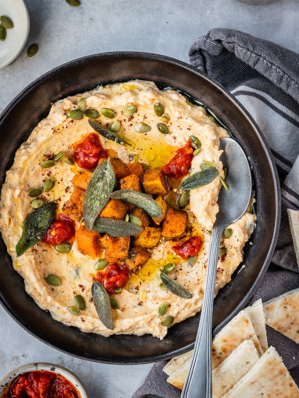 A bowl with mafghoussa sip garnished with roasted squash, pumpkin seeds, fried sage, sumac and chili paste.