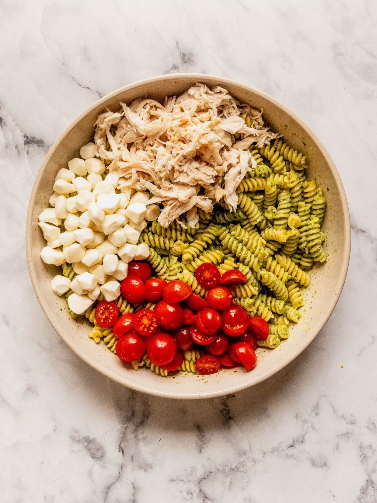 Large bowl with pasta tossed in pesto, layered with shredded chicken, mozzarella balls and cherry tomato slices.