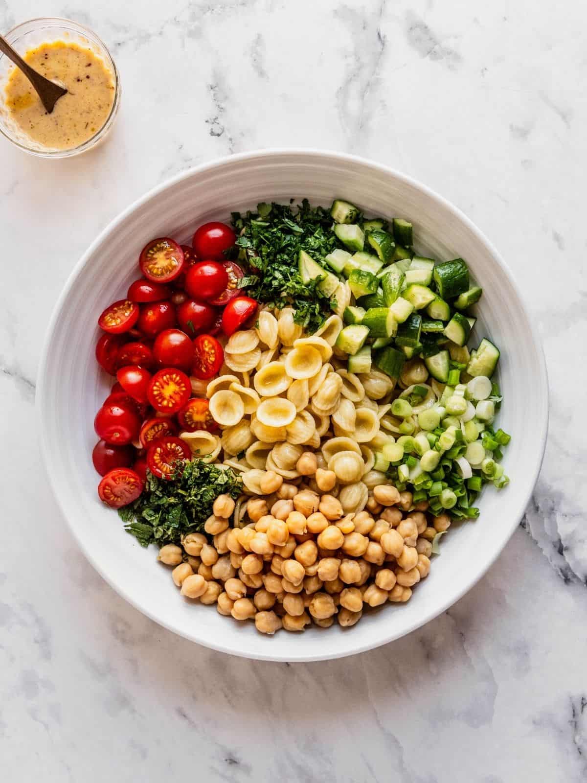 Large bowl with chopped cucumbers, halved cherry tomatoes, cooked pasta, chickpeas, and herbs.