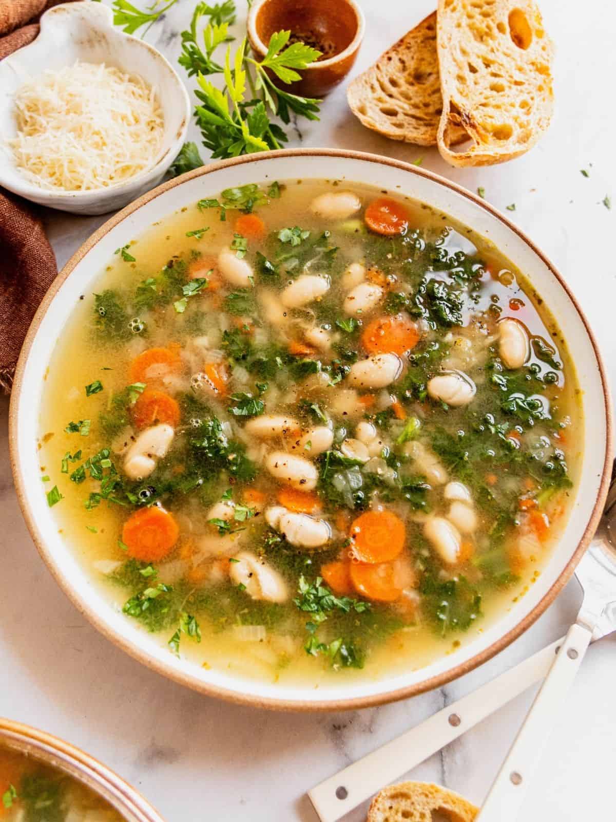 Tuscan white bean soup with kale in a bowl with shaved parmesan, sliced bread and herbs on the side.