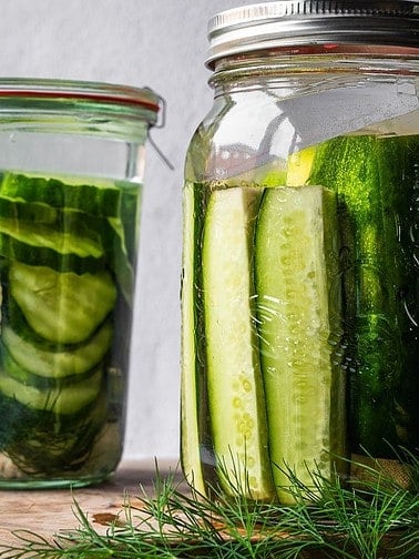 Two mason jars filled with sliced cucumbers with pickling brine and spices.