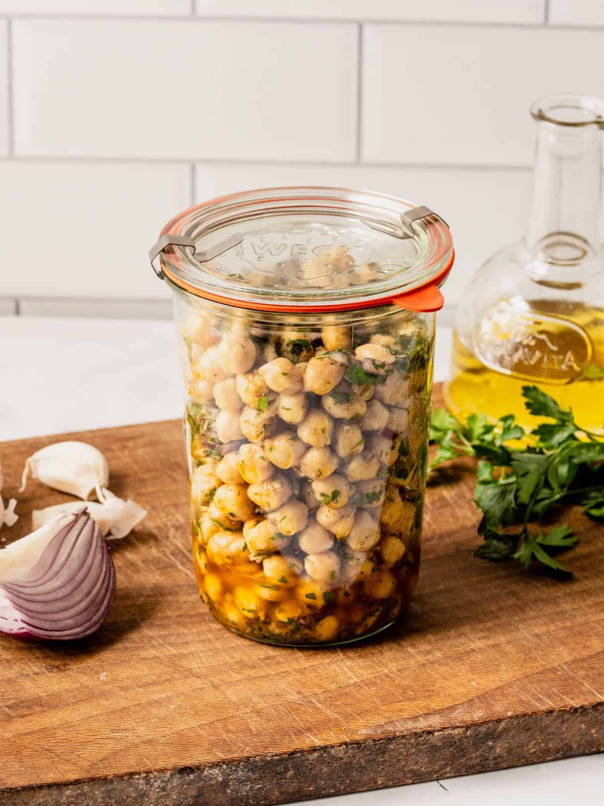 Marinated chickpeas in a mason jar with herbs, oil and seasoning sitting on a wooden board with olive oil, garlic and onion slices.