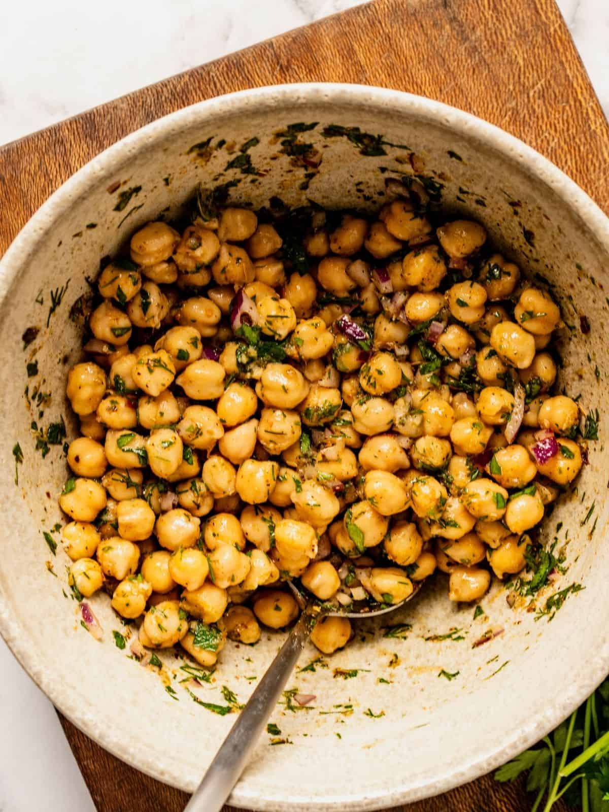 Marinated chickpeas in a bowl with herbs, oil and seasoning sitting on a wooden board.