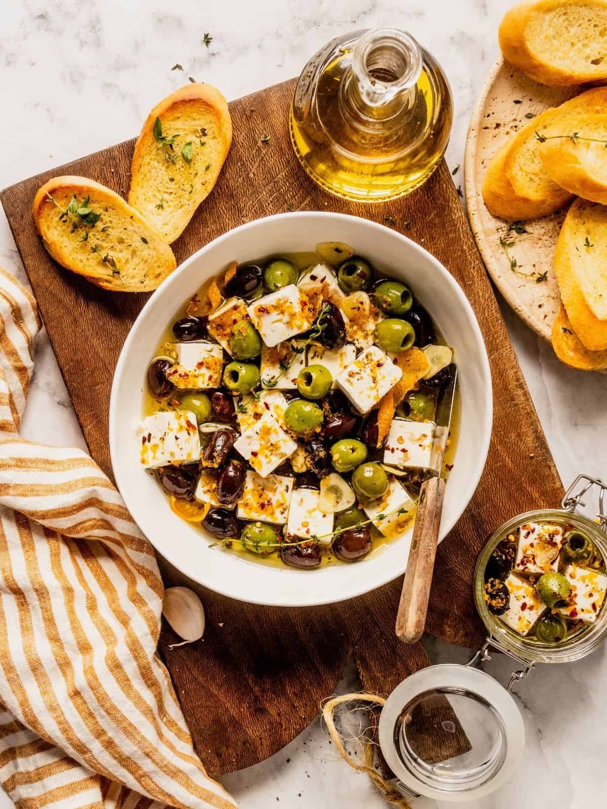 Citrus and herb marinated feta and olives in a bowl with thyme, orange peels, and spices, with sliced bread on the side.