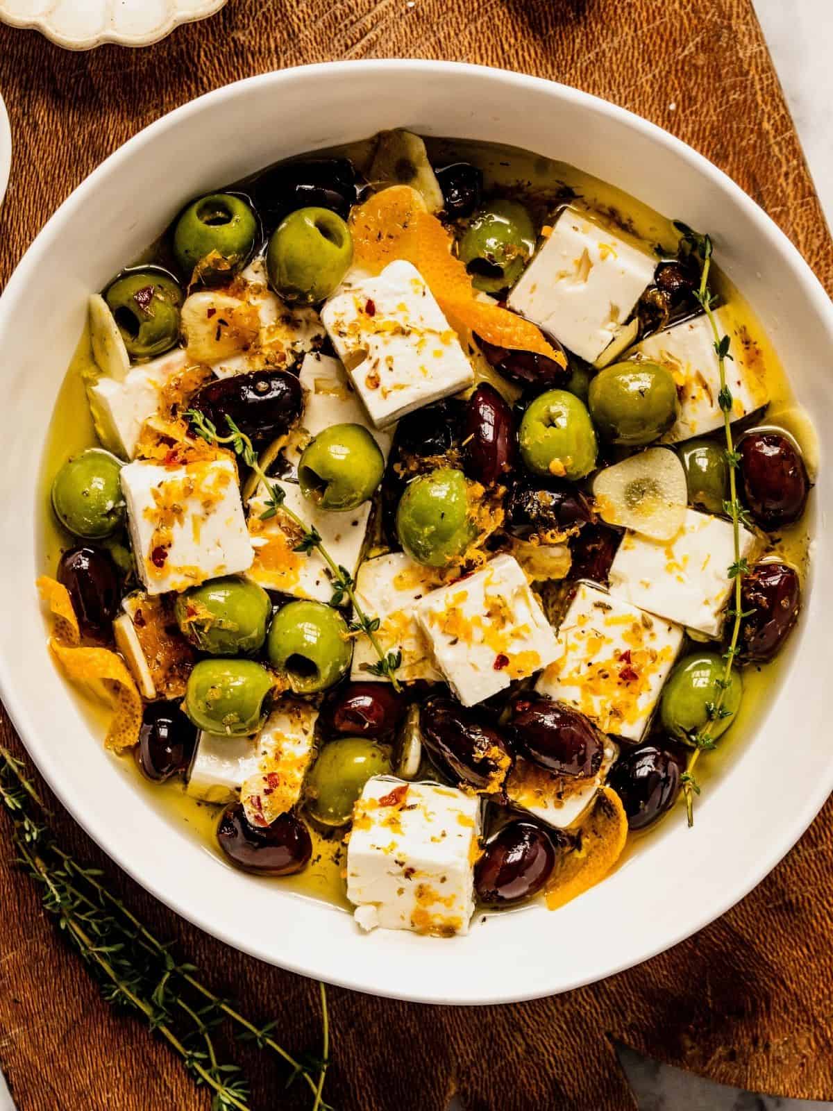 Citrus and herb marinated olives and feta in a bowl with thyme, orange peels, and spices.