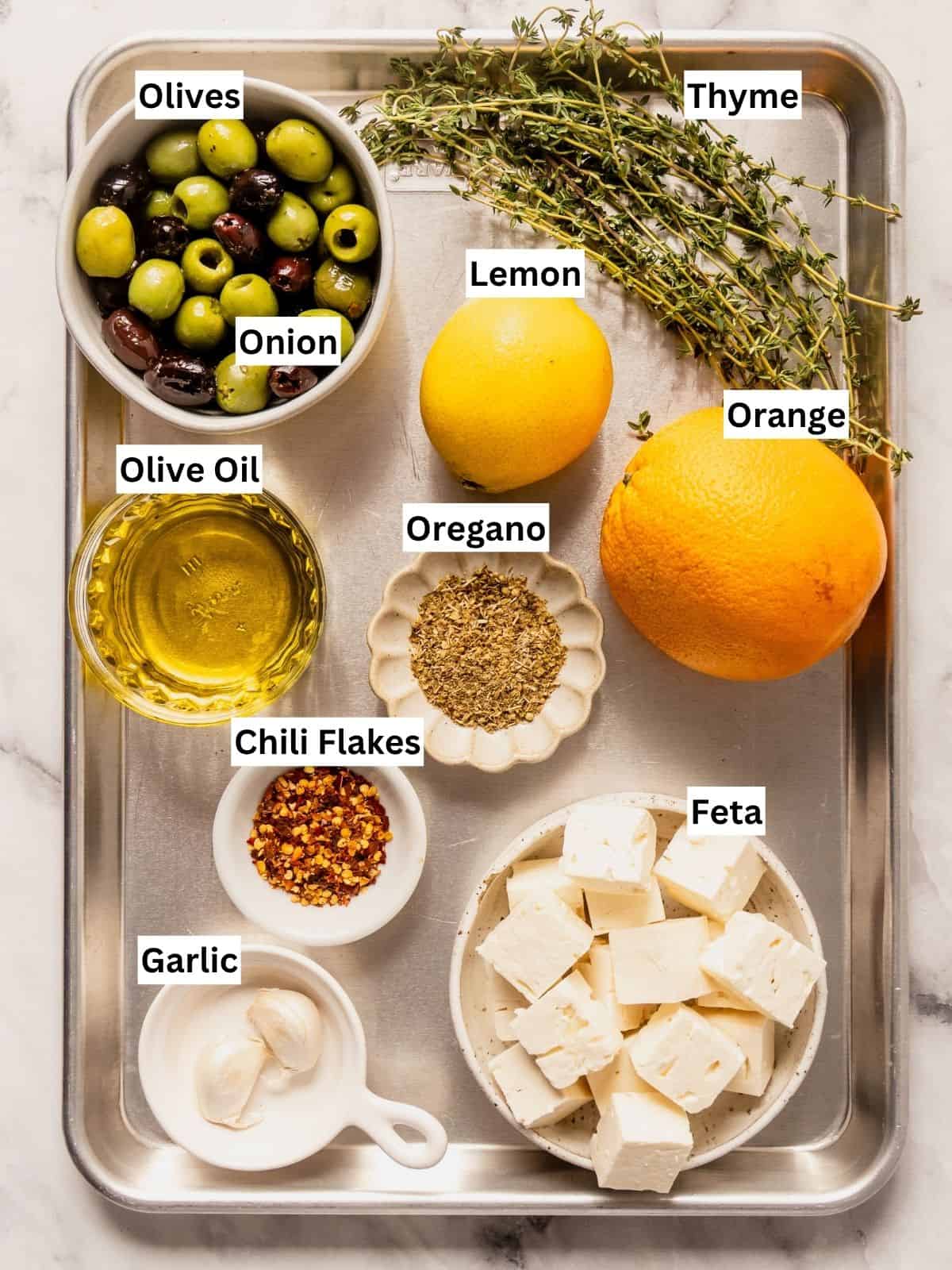Citrus and herb marinated feta and olives recipe ingredients with individual labels on a metal tray.