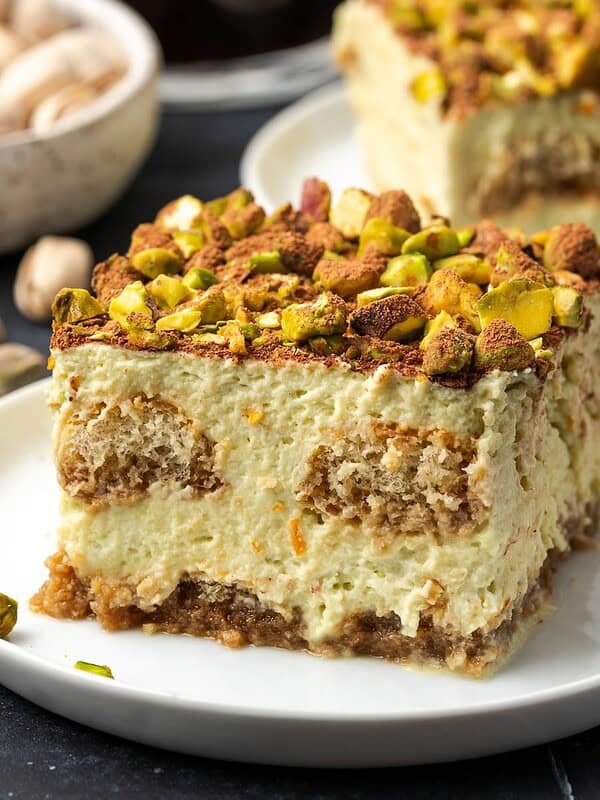 A slice of orange pistachio tiramisu decorated with crushed pistachios and cocoa on a plate.