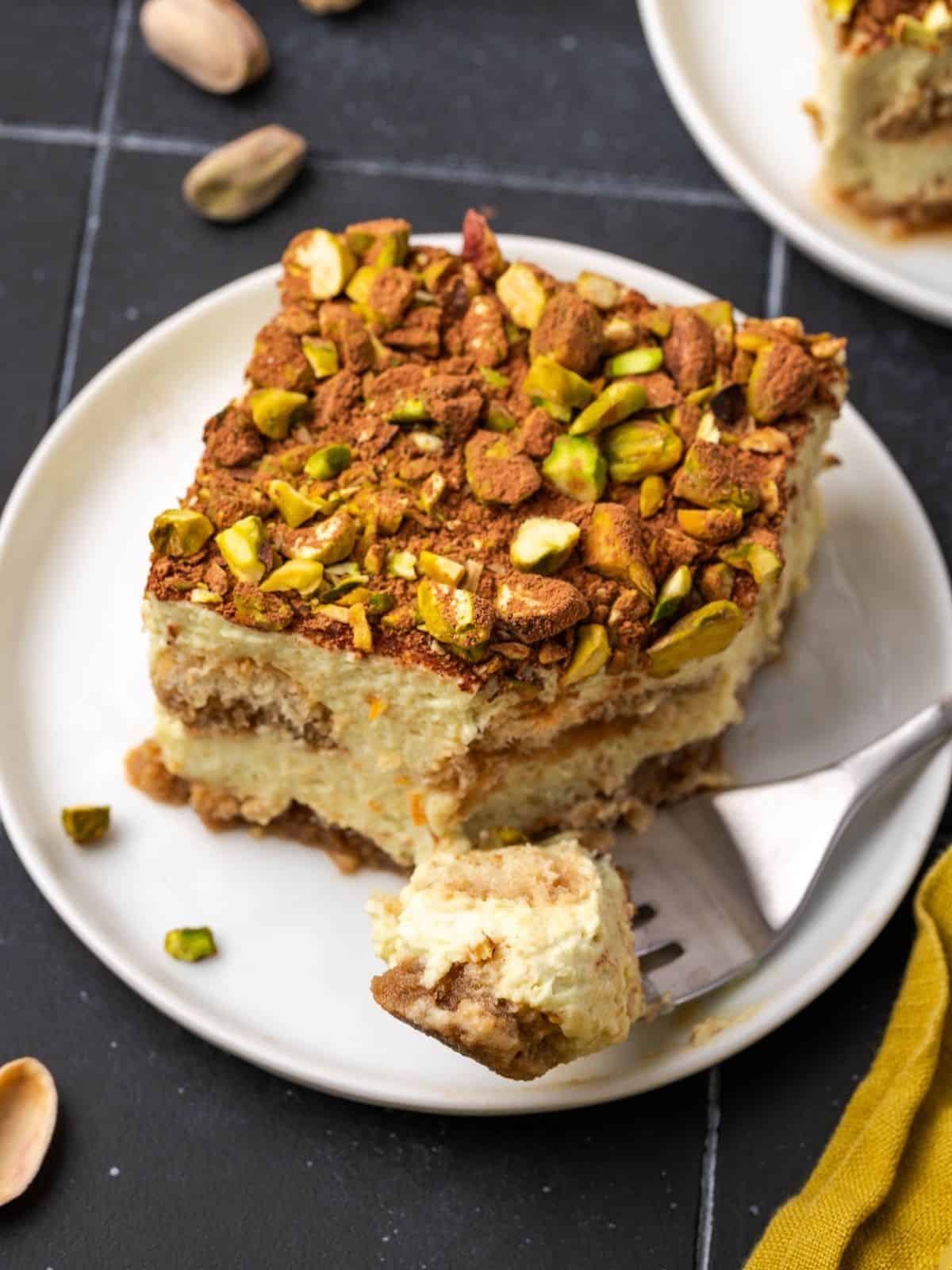 A slice of orange pistachio tiramisu decorated with crushed pistachios and cocoa on a plate.