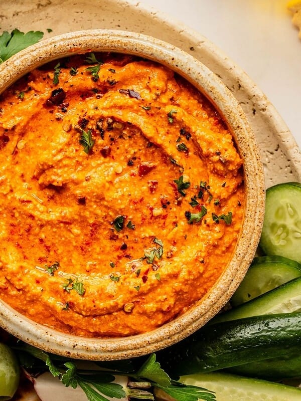 Bowl of red pepper feta dip (also known as Tirokafteri or Htipiti) on a plate garnished with parsley and served with bread, vegetables and olives.