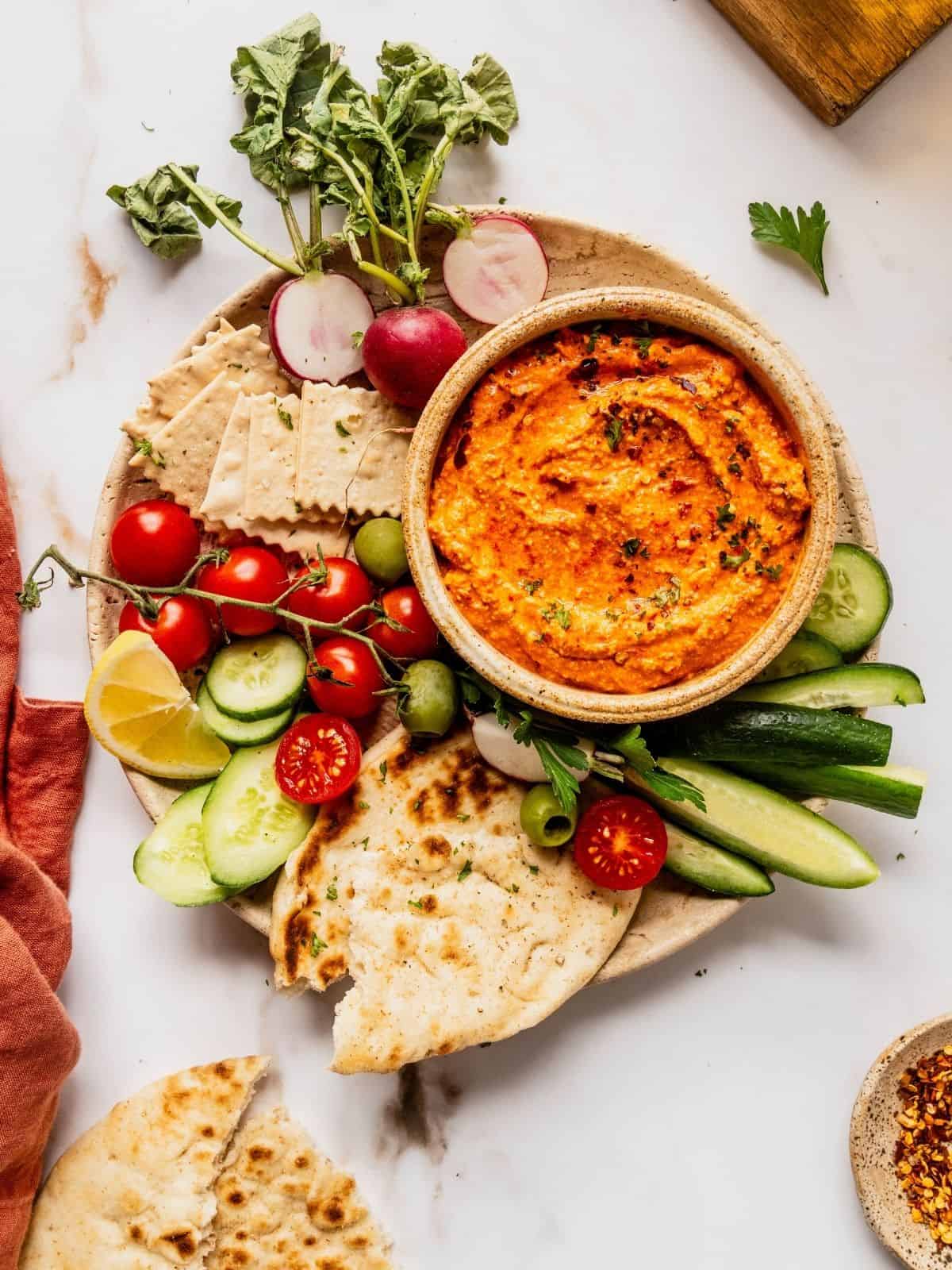 Bowl of red pepper feta dip (also known as Tirokafteri or Htipiti) on a plate garnished with parsley and served with bread, vegetables and olives.