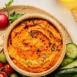 Bowl of red pepper feta dip (also known as Tirokafteri or Htipiti) on a plate garnished with parsley and served with bread, vegetables and olives.