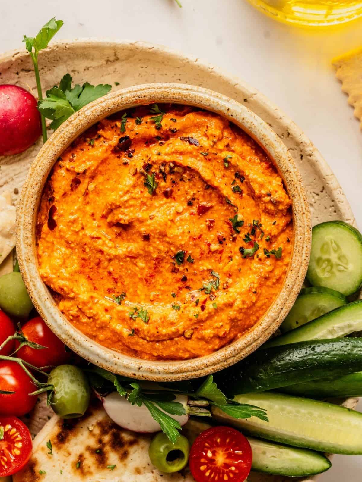 Bowl of red pepper feta dip (also known as Tirokafteri or Htipiti) on a plate garnished with parsley and served with bread, vegetables and olives.