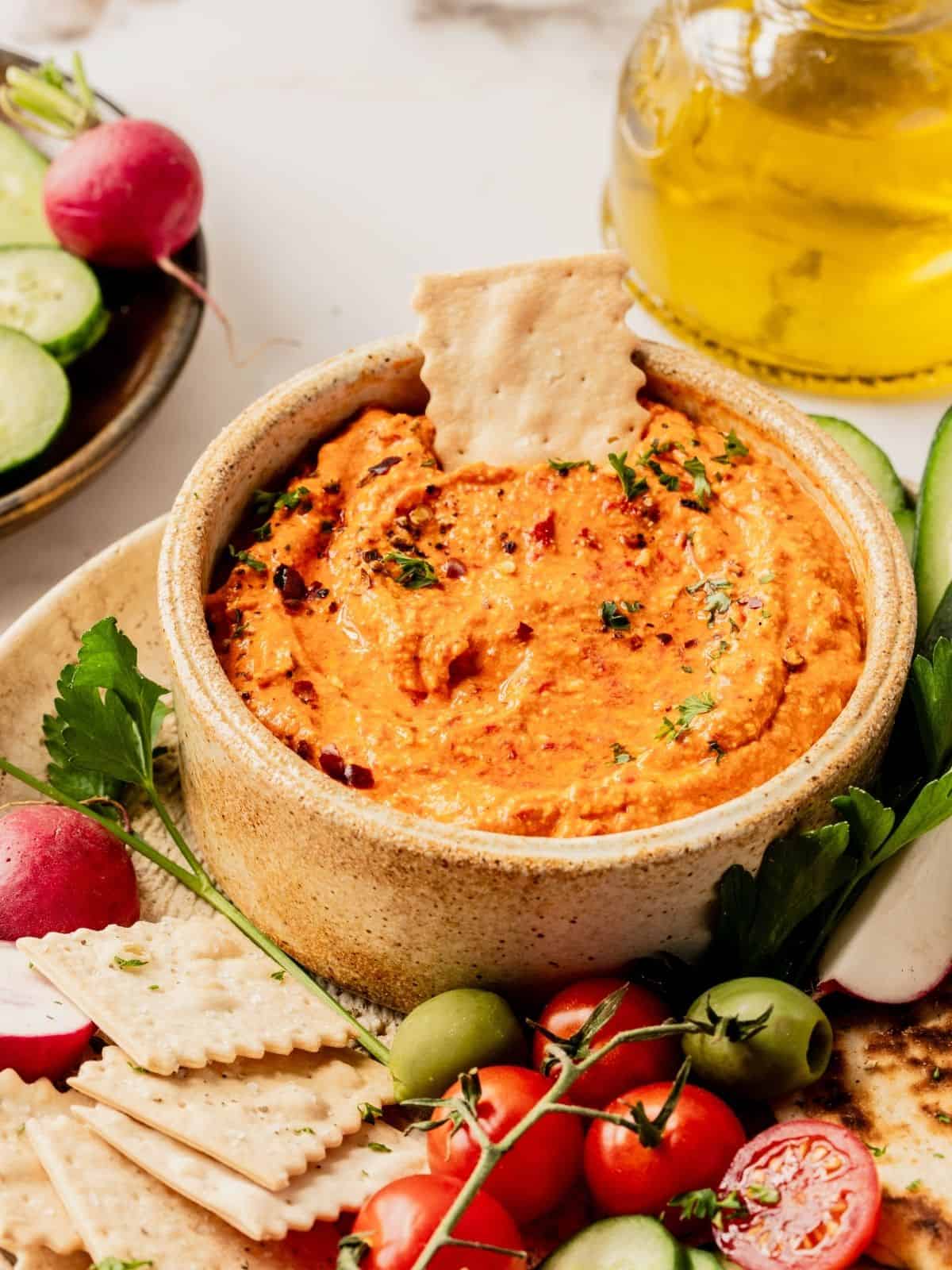 Bowl of red pepper feta dip (also known as Tirokafteri or Htipiti) on a plate garnished with parsley and served with bread, vegetables and olives.