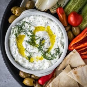 Bowl of creamy yogurt and feta dip, garnished with dill, with chopped vegetables and pita on the side.